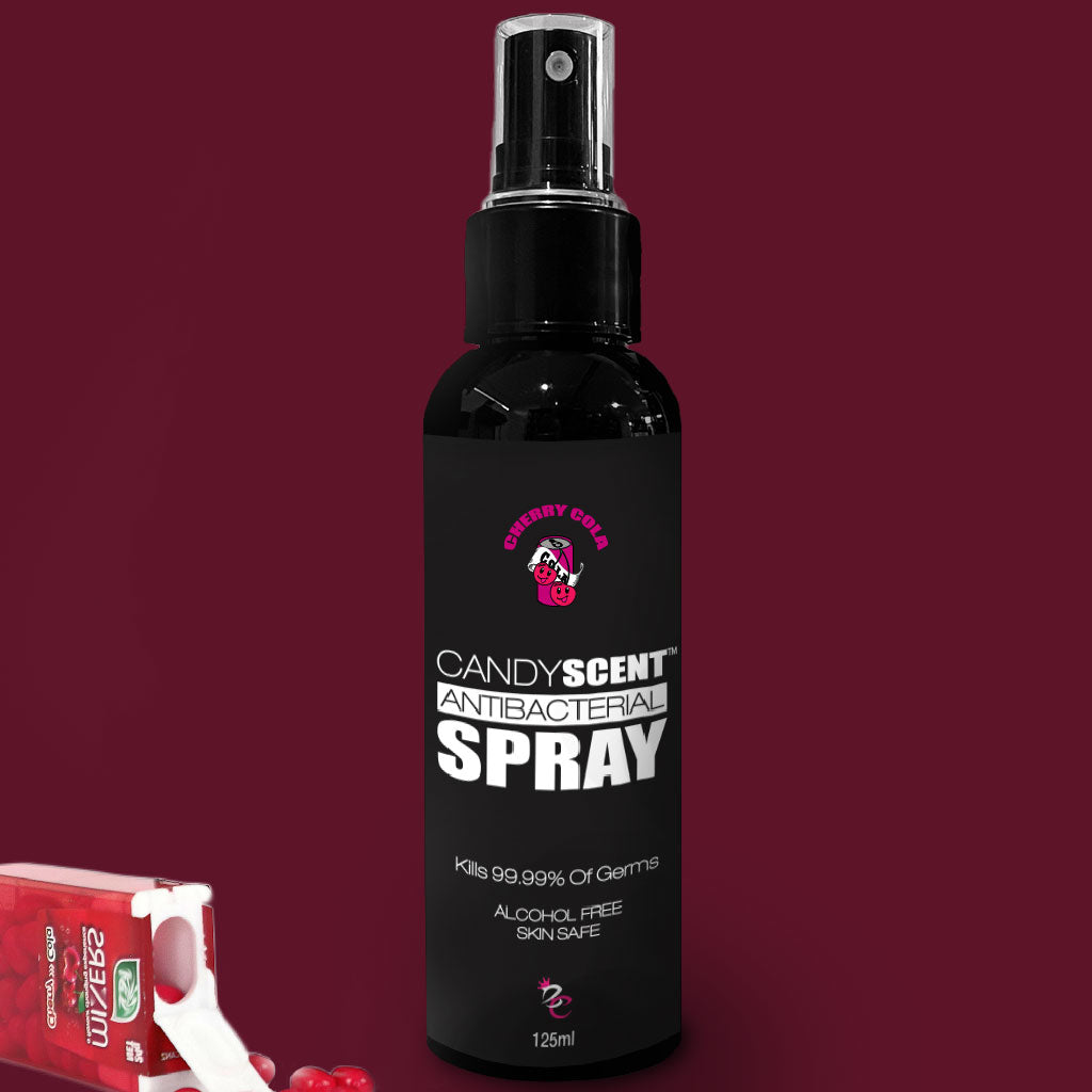A black bottle of CANDYSCENT™ Antibacterial Spray with a cherry cola scent sits against a reddish-burgundy background. The label highlights it is alcohol-free and kills 99.99% of germs; the bottle is 125 ml. To the left, a small overturned box with cherries is visible.