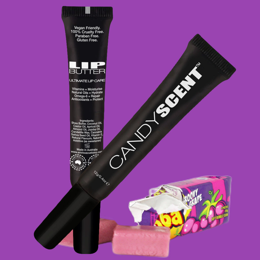 Two tubes of CANDYSCENT™ Lip Butter are angled against a purple background. One features the brand name "CANDYSCENT™," while the other displays "LIP BUTTER." In the foreground, colorful candy wrappers and pieces, including Bazooka bubble gum, add a playful touch.