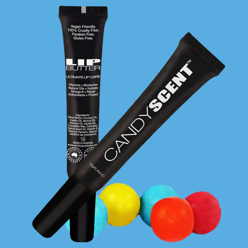 Two black tubes of moisturizing Lip Butter, branded with "CANDYSCENT™", are set against a blue background. The Lip Butter tube features labels declaring "Vegan Friendly, 100% Cruelty Free, Paraben Free, Gluten Free, made with natural ingredients." Four colorful round candies are artfully arranged below the tubes.