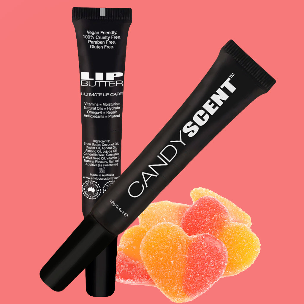 Two tubes of Lip Butter from the brand CANDYSCENT™ are displayed against a pink background with gummy heart candies. The tubes read "LIP BUTTER Ultimate Lip Care" and "CANDYSCENT." These moisturizing products, made with natural ingredients, are labeled as vegan, cruelty-free, paraben-free, and gluten-free.