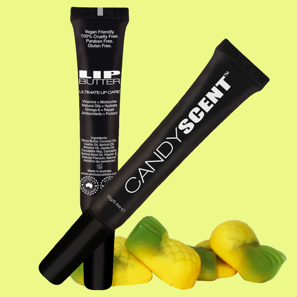 Two black tubes of lip care products, labeled "Lip Butter" by CANDYSCENT™, stand against a light yellow background, complemented by yellow and green gummy candies at their base. The moisturizing formula boasts natural ingredients for soft, hydrated lips.