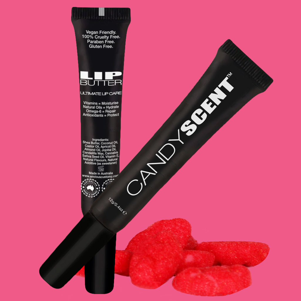 Two black tubes of lip balm are displayed against a pink background. One tube, branded "CANDYSCENT™ Lip Butter," stands upright, showcasing its moisturizing natural ingredients, while the other is tilted and leaning on the first. Red gummy candy hearts are positioned at the base of the tubes.