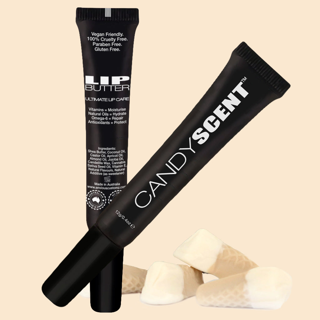 Two black tubes of Lip Butter from CANDYSCENT™ are displayed on a beige background. The tubes, labeled "Lip Butter" and showcasing the brand name "CANDYSCENT™," promise to be vegan, cruelty-free, paraben-free, and gluten-free. They are crafted with natural ingredients. Below them, several white wafer-style candies are arranged beautifully.