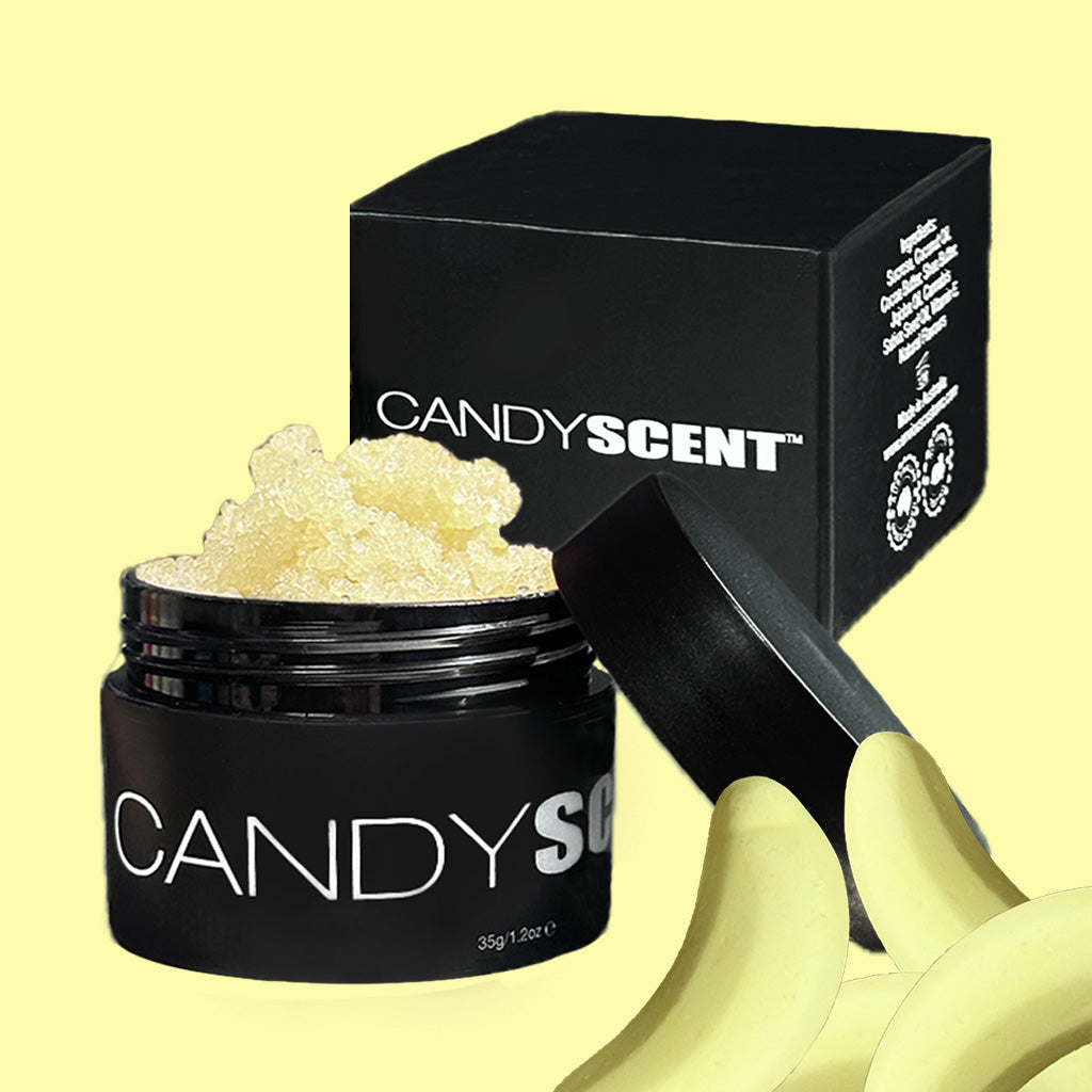 The image features an open black jar labeled "CANDYSCENT™" filled with a textured substance, which is likely their Lip Sugar Scrub designed for exfoliating dead skin. The jar is positioned next to a matching black box bearing the same label. Surrounding the scene are peeled banana slices on a yellow backdrop, highlighting the nourishing oils of the product.