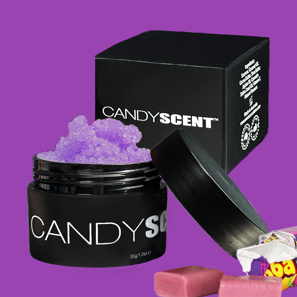 Open a black container labeled "CANDYSCENT™" filled with a purple crystal-like substance, presumably the Lip Sugar Scrub infused with nourishing oils, with the lid resting beside it. Behind it stands a matching black box bearing the same "CANDYSCENT™" label. In the foreground, wrapped candy pieces are scattered against a purple background.