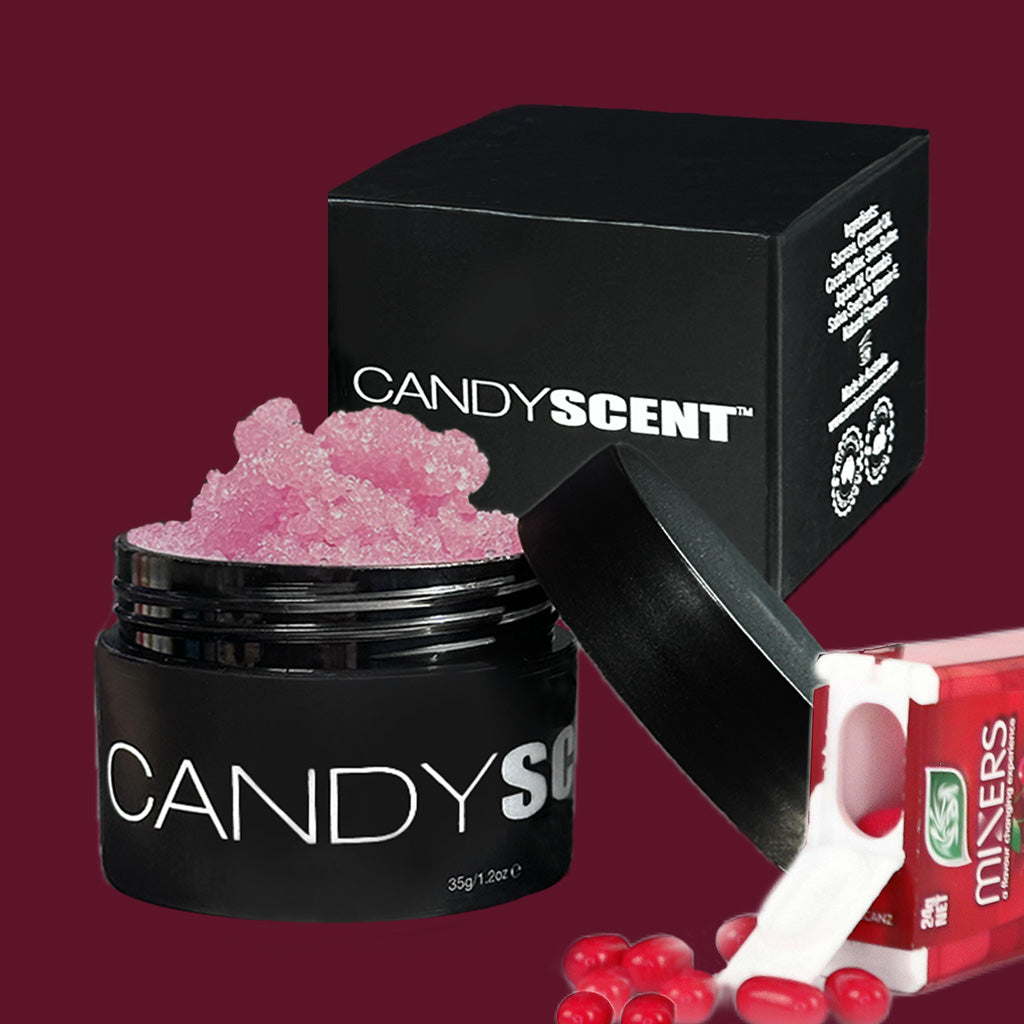 A jar of pink, candy-scented crystals with a black lid, labeled "CANDYSCENT™ Lip Sugar Scrub," sits open next to its black packaging box. The background shows several small red candies and another candy package against a solid maroon color. Infused with nourishing oils, these Lip Sugar Scrubs exfoliate dead skin effectively.