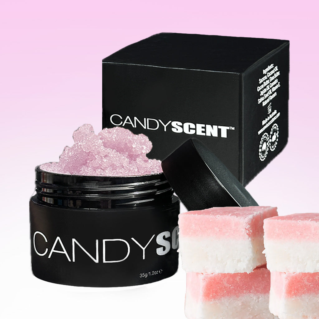 An open black container labeled "CANDYSCENT™" reveals pink crystal-like contents. Behind it sits a matching black box with the CANDYSCENT™ branding. In the foreground, square, layered pink and white Lip Sugar Scrubs that exfoliate dead skin are displayed. The background transitions from white to light pink in a gradient pattern.