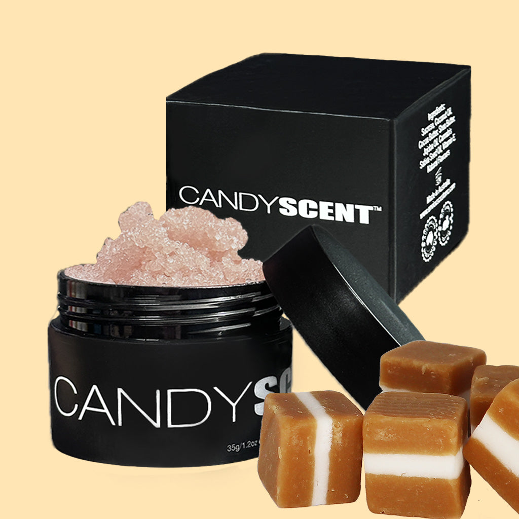 A black container labeled "CANDYSCENT™ Lip Sugar Scrub" is open, revealing a pink granulated substance inside. Beside it is a matching black box with the same branding. In the foreground, there are several caramel-colored squares with white layers. This product is perfect for exfoliating dead skin and is infused with nourishing oils.