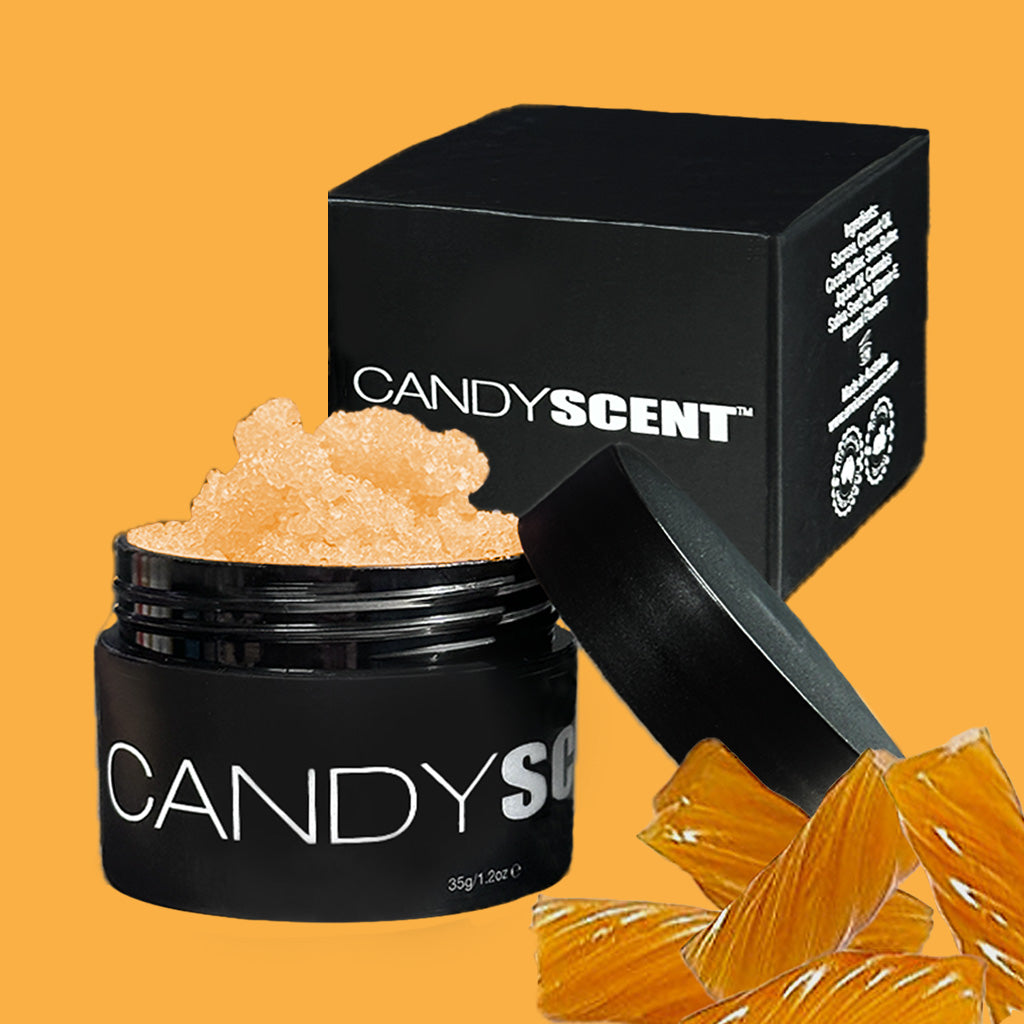A black jar labeled "CANDYSCENT™ Lip Sugar Scrub" contains an orange, crystallized substance. Behind it, a coordinating black box features white text. In the foreground, amber-colored rock candy-like pieces are placed on the right against an orange background, suggesting that this product exfoliates dead skin and nourishes your lips.