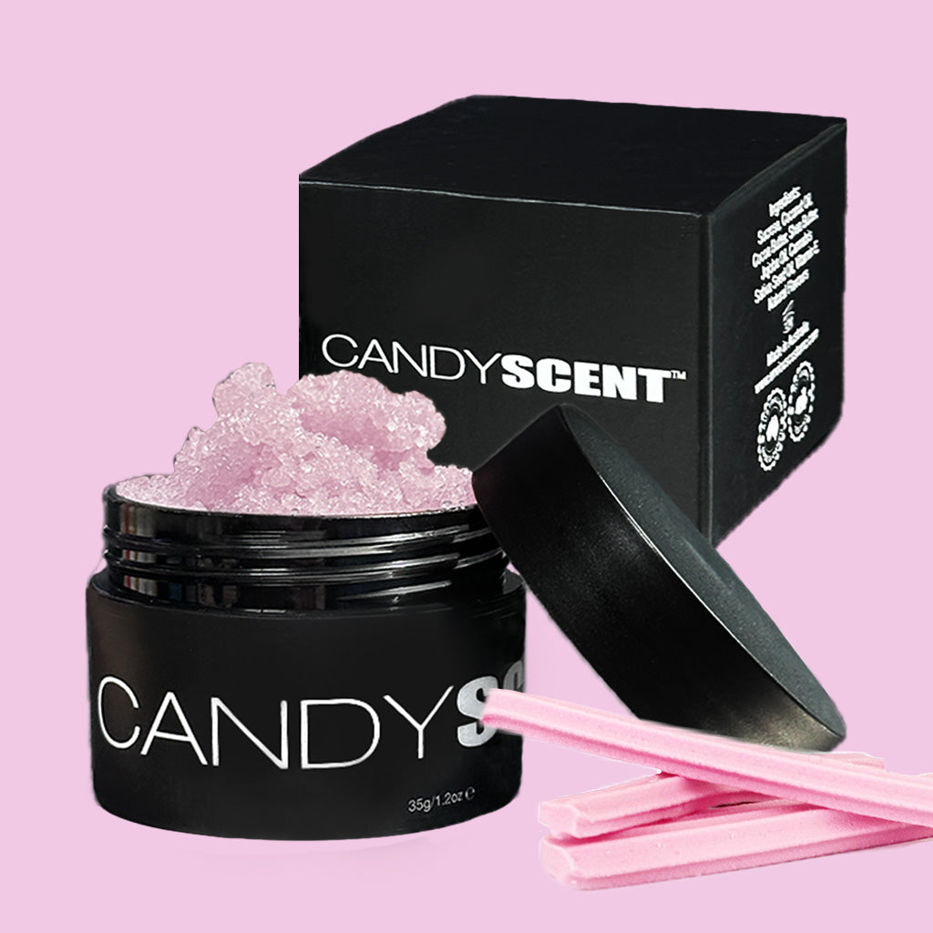 A black container labeled "CANDYSCENT™ Lip Sugar Scrub" is filled with light pink crystal-like granules and sits next to its black, partially open lid. Behind it, a black box with the same label is displayed. Three pink sticks are placed in the foreground on a pink background.
