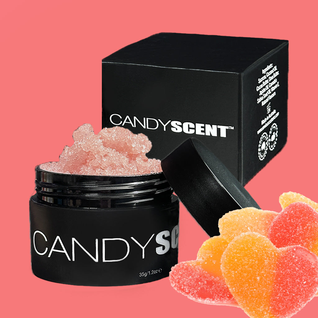A black container branded with "CANDYSCENT™" and filled with pink sugar granules is displayed in front of a matching black box with white text. Nearby, heart-shaped gummy candies in red and peach shades are also shown. The background is solid pink, echoing the nourishing oils in this Lip Sugar Scrub designed to exfoliate dead skin.