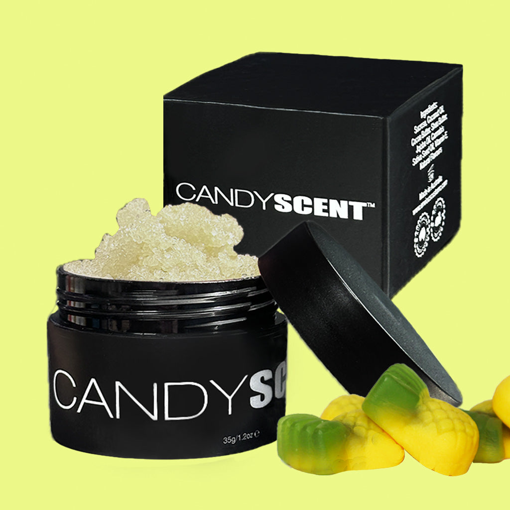 A black jar of CANDYSCENT™ Lip Sugar Scrub with its lid resting to the side, next to a black box and some yellow and green candy-shaped objects, all set against a light green background. Infused with nourishing oils, this scrub helps exfoliate dead skin for soft, smooth lips.