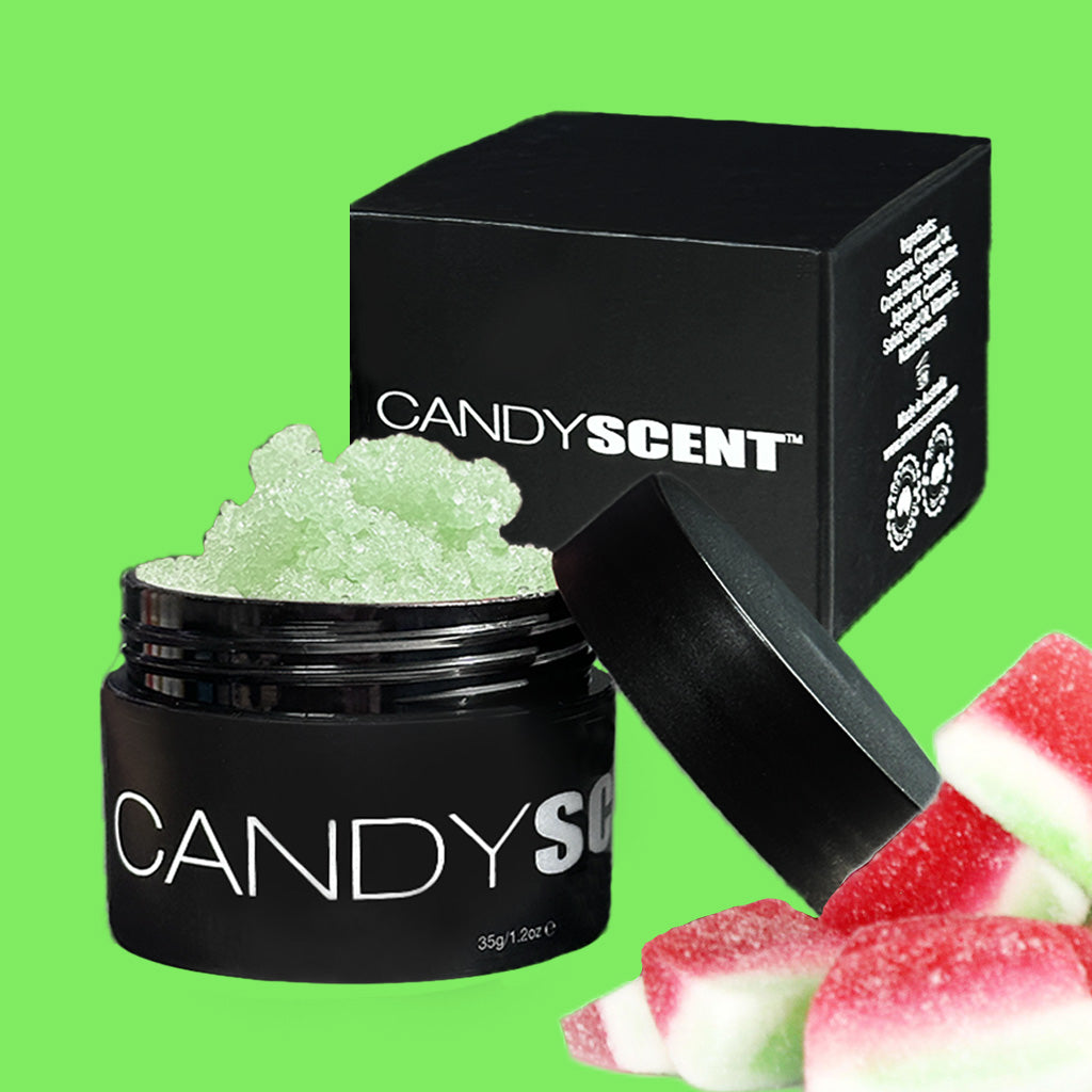 A jar of CANDYSCENT™ Lip Sugar Scrub with its lid removed, revealing a green, grainy substance inside. The open jar is placed in front of its black box packaging, with slices of red and white candy positioned next to it on a bright green background. Perfect for exfoliating dead skin and packed with nourishing oils.