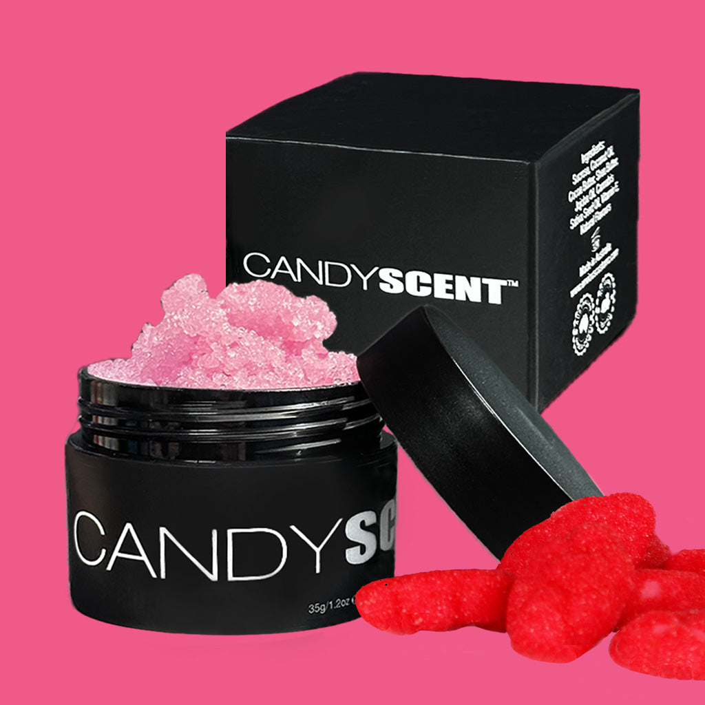 A black jar labeled "CANDYSCENT™" with its lid off reveals pink, textured Lip Sugar Scrubs. The jar sits in front of a black box with the same branding. Red gummy candies are positioned to the right of the jar and box, all set against a pink background.