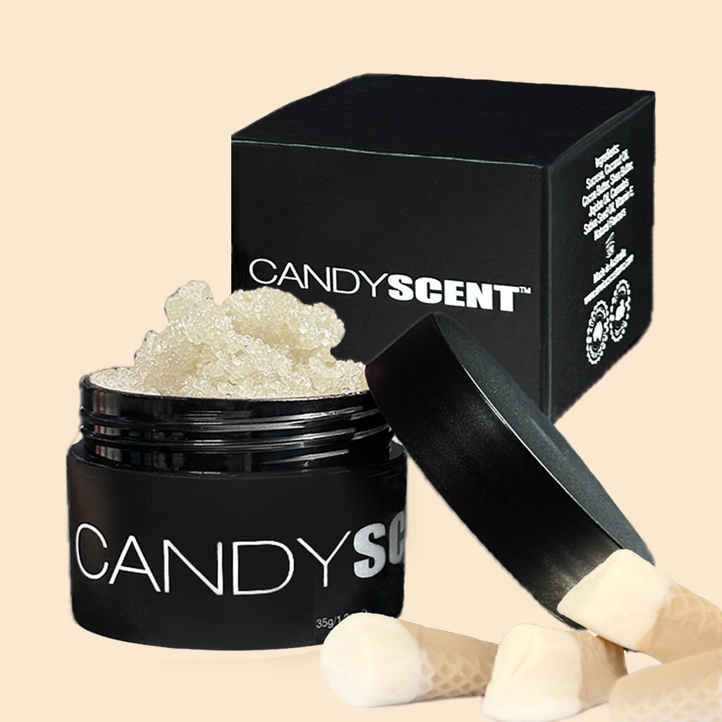 A black container labeled "CANDYSCENT™ Lip Sugar Scrub" with a white, grainy substance inside rests with its lid partially open. Behind the container is a black box that reads "CANDYSCENT™" and shows details. Several cone-shaped, beige objects lie in the foreground, hinting at nourishing oils in these Lip Sugar Scrubs.