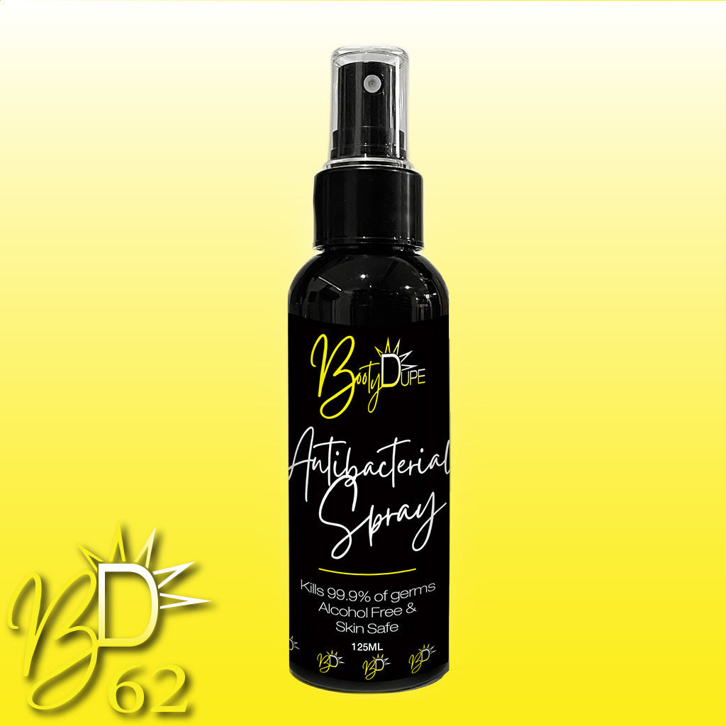 A 125ml bottle of "BOOTY DUPE Antibacterial Spray" is centered on a gradient yellow background. The black bottle features white text and a spray nozzle. The label highlights that the alcohol-free spray kills 99.99% of germs and is safe for skin. The logo "BD62" is in the bottom left corner.