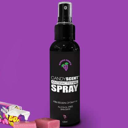 A black bottle labeled "CANDYSCENT™ Antibacterial Spray" with a green cartoon character and "Grape" text prominently displayed, set against a vibrant purple background. The antibacterial spray boasts the ability to kill 99.99% of germs, is free from alcohol, and safe for skin use. Surrounding the bottle are a few colorful candies.