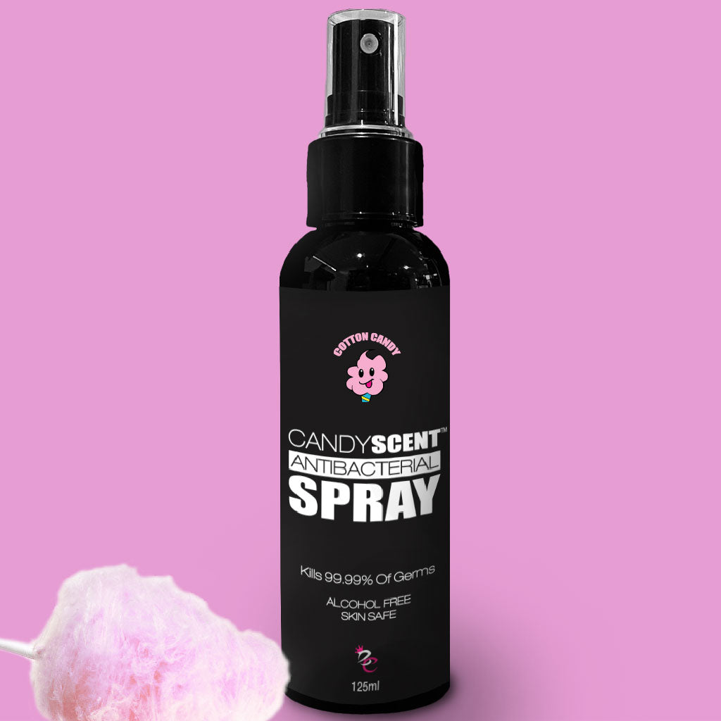 A black bottle of Antibacterial Spray by CANDYSCENT™ features a pink cotton candy graphic on a pink background. The text on the bottle highlights that it kills 99.99% of germs, is alcohol-free, and skin-safe. A cotton candy prop is positioned at the bottom left corner.