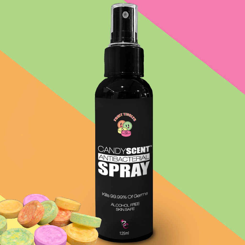 A 125ml bottle of CANDYSCENT™ Antibacterial Spray features a black label adorned with a cartoon candy character. This alcohol-free spray claims to kill 99.99% of germs and is safe for skin use. The background displays diagonal stripes in vibrant yellow, green, orange, and pink hues.