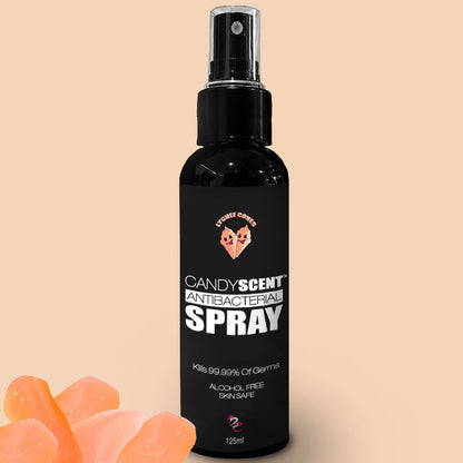 A black spray bottle labeled "Antibacterial Spray" by CANDYSCENT™ with a tagline "Kills 99.99% of Germs" sits against a beige background. The alcohol-free, 125ml bottle is featured alongside orange translucent rocks in the bottom left corner of the image.