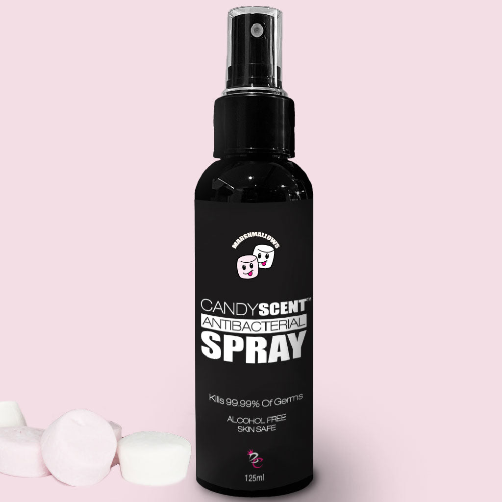 A black bottle labeled "CANDYSCENT™ Antibacterial Spray" with a pump top on a pink background. The bottle text states it kills 99.99% of germs and is alcohol-free and skin-safe. A few white and pink marshmallows are placed next to the antibacterial spray.