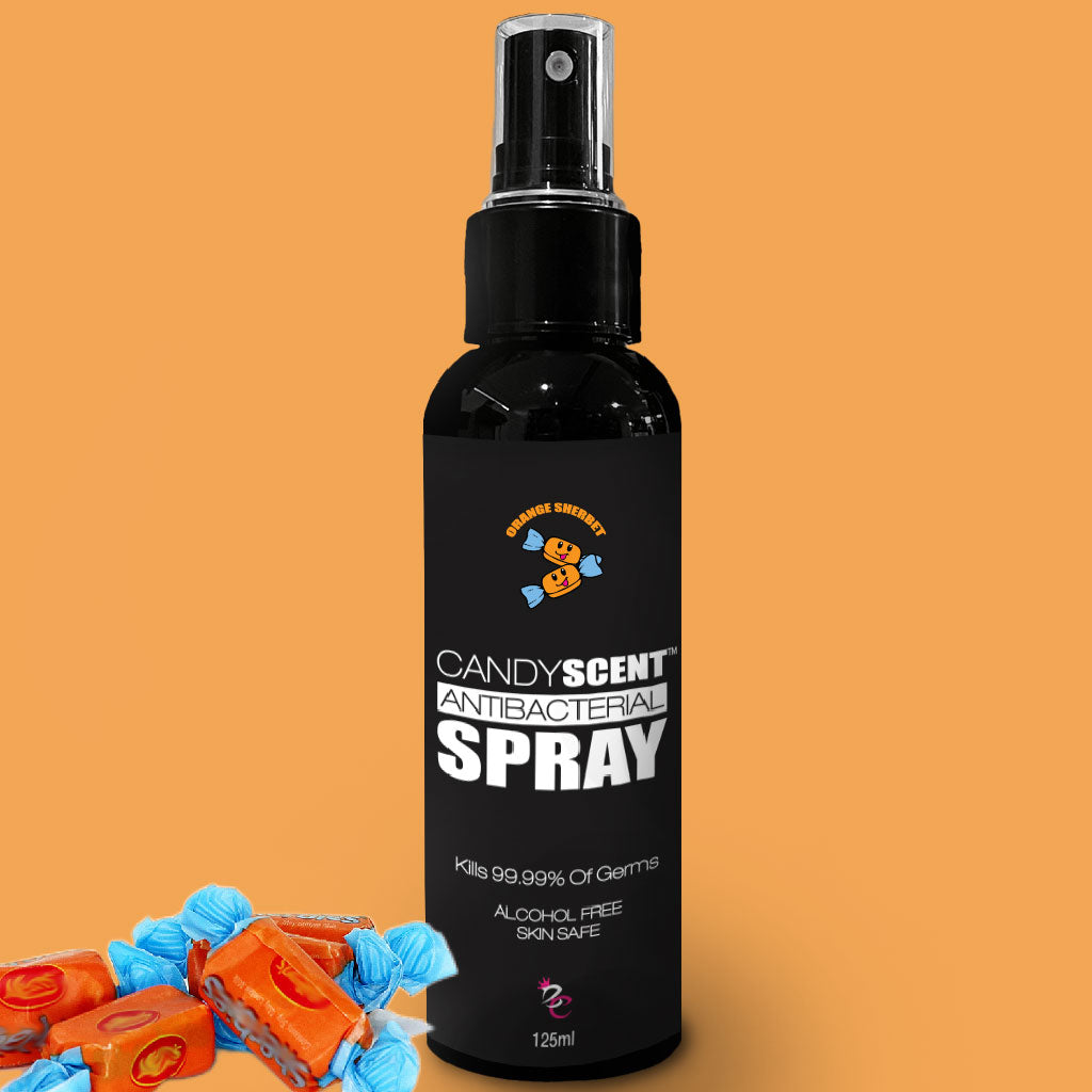 A black spray bottle labeled "CANDYSCENT™ Antibacterial Spray," promising to kill 99.99% of germs without the use of alcohol, ensuring it's skin-safe. The label features a cartoon bear, while small piles of orange and blue wrapped candies are positioned around the bottle.