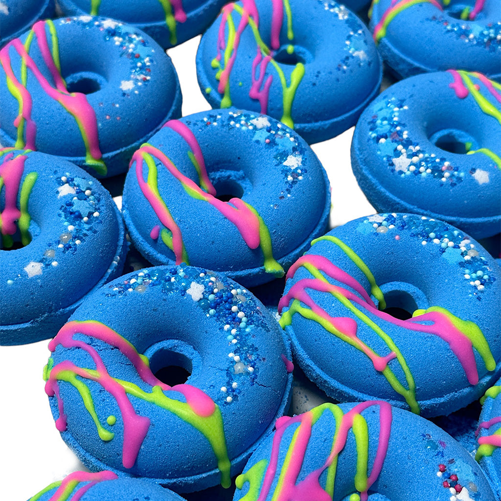 A group of vibrant blue donuts decorated with colorful drizzles of pink and green icing, and sprinkled with white stars and blue sugar crystals. Reminiscent of CANDYSCENT™ Bath Bombs, these treats are neatly arranged on a white background.