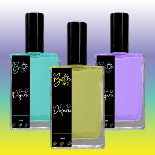 Image of three perfume bottles with black caps. The bottles feature colorful liquids: teal, green, and purple, each with a "BOOTY DUPE" branded label displaying different numbers (39, 62, 84) and the text "Eau De Perfume." These long-lasting tropical fragrances are set against a gradient background of blue to yellow.