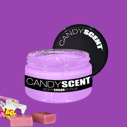 A jar of CANDYSCENT™ Body Sugar Scrub, featuring a purple scrub inside, is placed on a purple background. The jar is adorned with a black label and white text. To the left of the jar lies a partly unwrapped candy bar, surrounded by pink pieces, highlighting its promise to exfoliate and hydrate using nourishing oils.