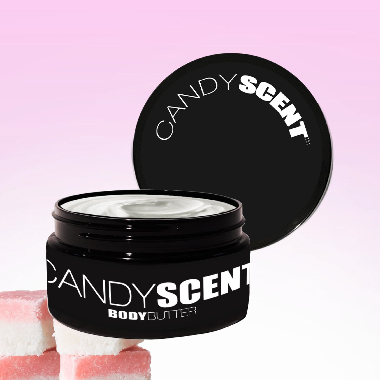 An open jar of CANDYSCENT™ Body Butter, topped with a black lid, reveals its creamy texture amidst a pink gradient backdrop, offering promises of hydration and nourishment. Complementing the scene are three pink and white cube-shaped items placed below, enhancing the delightful display.