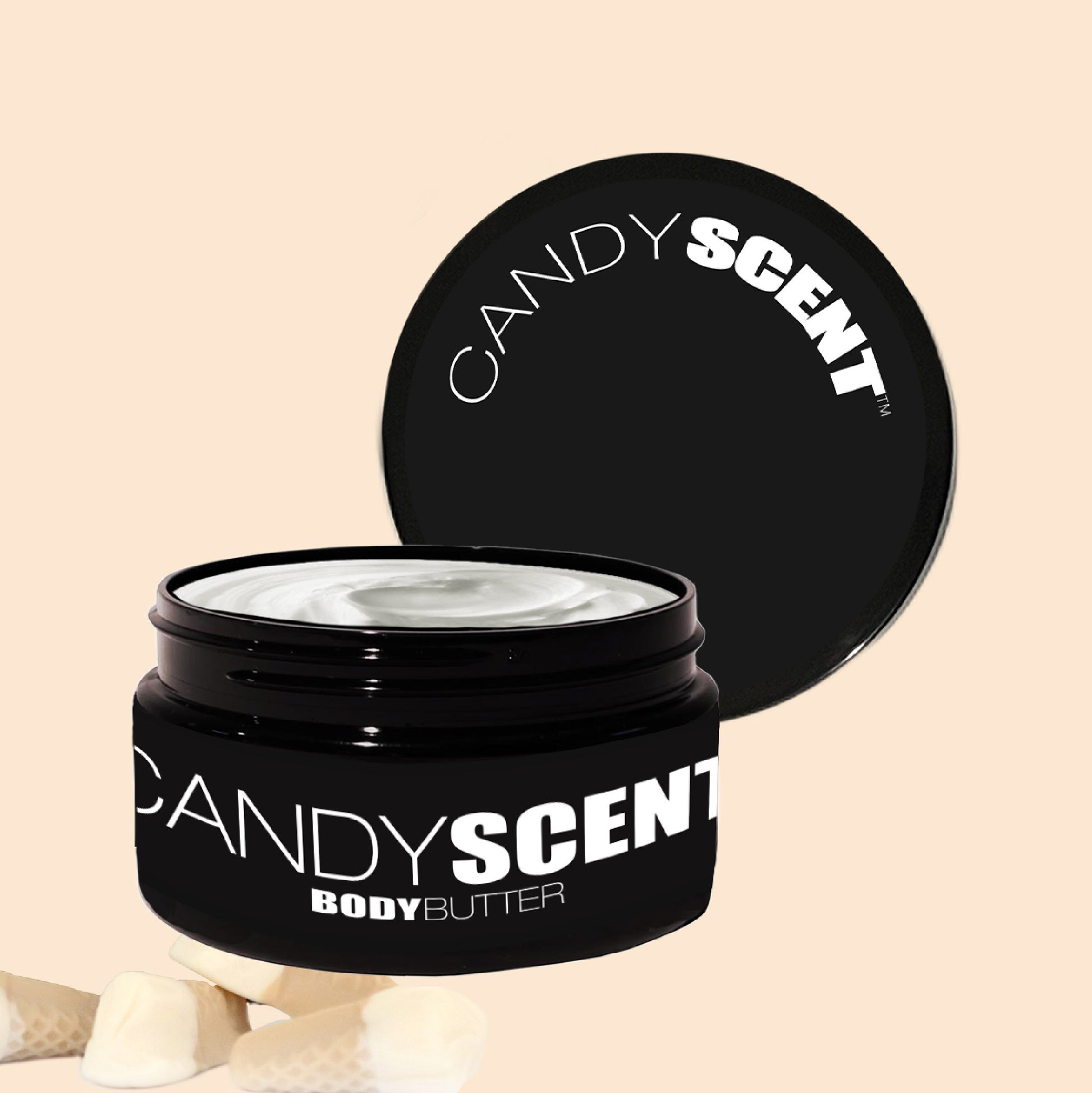 The CANDYSCENT™ Body Butter jar is open, displaying a creamy white texture that ensures hydration and nourishment. The black lid, featuring bold white text, rests against the jar. A soft beige background highlights small white candy pieces at the bottom.