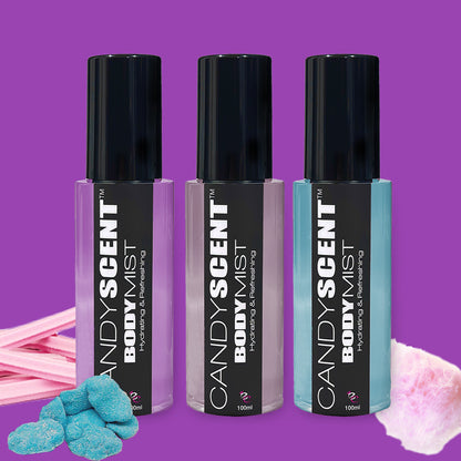 The image showcases three bottles of CANDYSCENT™ Hydrating Body Mist, each uniquely colored in black, pink, and light blue. These bottles are set against a vibrant purple background adorned with cotton candy and various candy pieces, emphasizing the nourishing ingredients such as witch hazel.