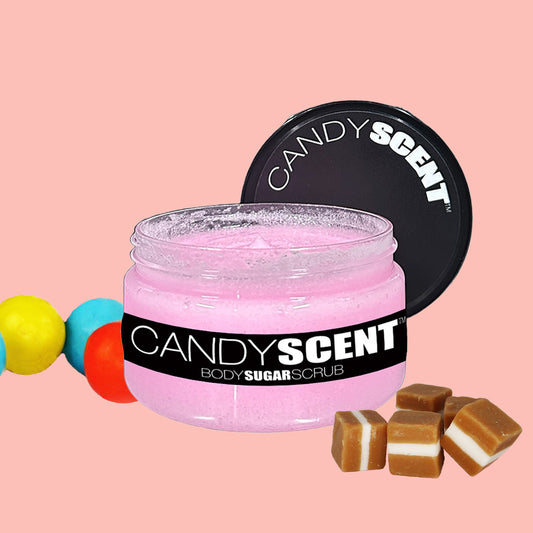 A jar of CANDYSCENT™ Body Sugar Scrub with a black lid, containing pink scrub. Colorful spheres and caramel candy pieces are placed on either side of the jar, all set against a pink background. Infused with nourishing oils, it promises to exfoliate and hydrate your skin for a glowing finish.