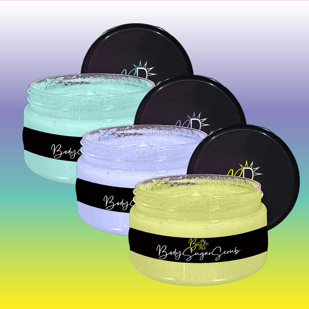 Three jars of BOOTY DUPE Body Sugar Scrub in pastel colors (mint green, lavender, and lemon yellow) are displayed with their lids partially open. Each jar features a black label with decorative text and design. Perfect for all skin types, these scrubs gently exfoliate and hydrate. The background is a gradient of blue to yellow.