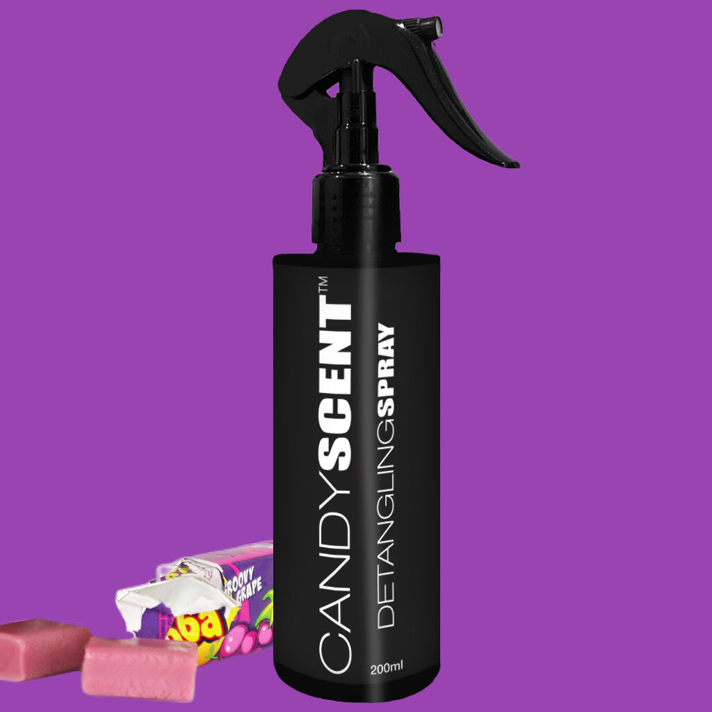 A black spray bottle labeled "CANDYSCENT™ Hair Detangling Spray 200ml" is set against a purple background. Surrounding the bottle, unwrapped and wrapped pieces of pink candy enhance the candy theme. Infused with Aloe Vera, this spray works wonders on all hair types.