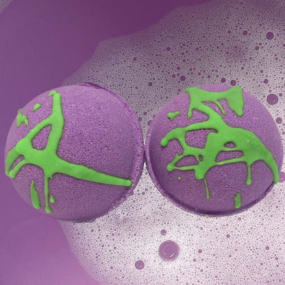 Two round purple Bath Bombs by CANDYSCENT™, adorned with green drizzles, float in a tub of purple-colored water topped with a layer of foam. As they dissolve, they release sweet scents and are crafted from natural ingredients.