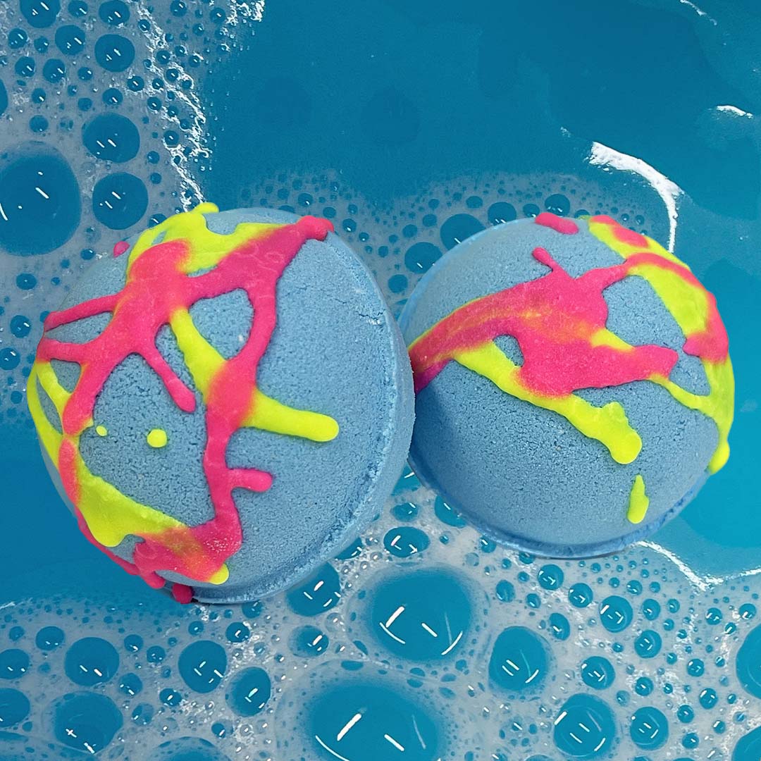 Two CANDYSCENT™ Bath Bombs, featuring vibrant pink and yellow splashes against a blue backdrop, rest on a bubbly surface in blue water, filling the air with sweet aromas and crafted from natural ingredients.