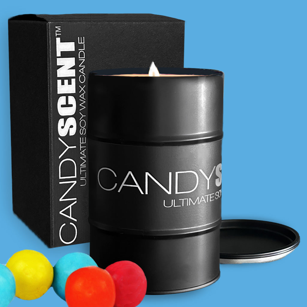 A lit black soy wax candle tin from the CANDYSCENT™ brand is partially opened, with the lid resting beside it. The candle sits in front of its sleek black packaging box on a blue background, accompanied by three vibrant gumballs in yellow, red, and blue.