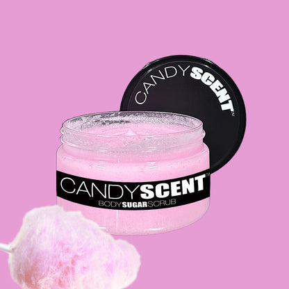 A jar of CANDYSCENT™ Body Sugar Scrub with a black lid is open, displaying the pink scrub within. The jar's label features a black background with white text. Positioned in the foreground is an image of pink cotton candy, while the solid pink background highlights the nourishing oils that exfoliate and hydrate.