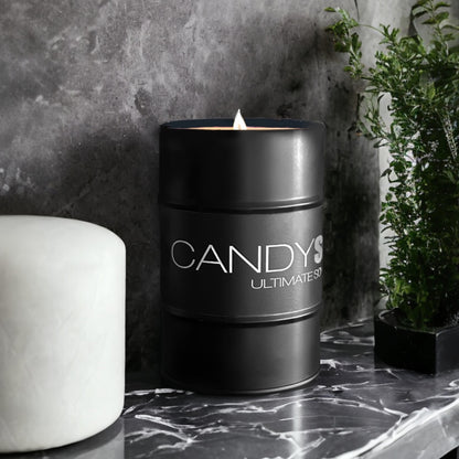 A black cylindrical Soy Wax Candle Tin from CANDYSCENT™ sits on a glossy, dark marble surface. The candle flame is lit, casting a warm glow. To the left is a white cylindrical object, and to the right, a small green plant adds a touch of nature. Enjoy the ambiance with CANDYSCENT™ candles.