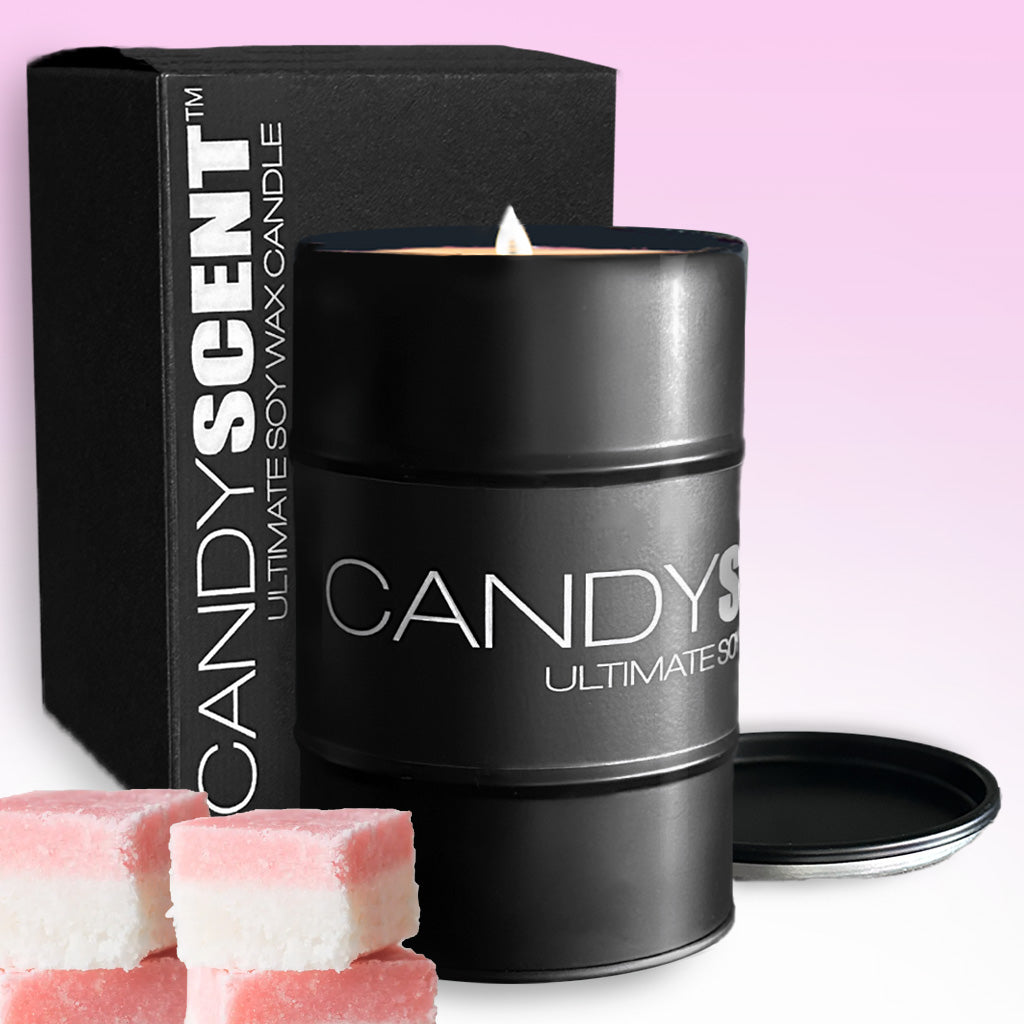 A lit black cylindrical soy wax candle tin bearing the "CANDYSCENT™" brand, positioned in front of its matching black box adorned with white text. In the foreground, pink and white square confections are set against a subtle pink gradient background.