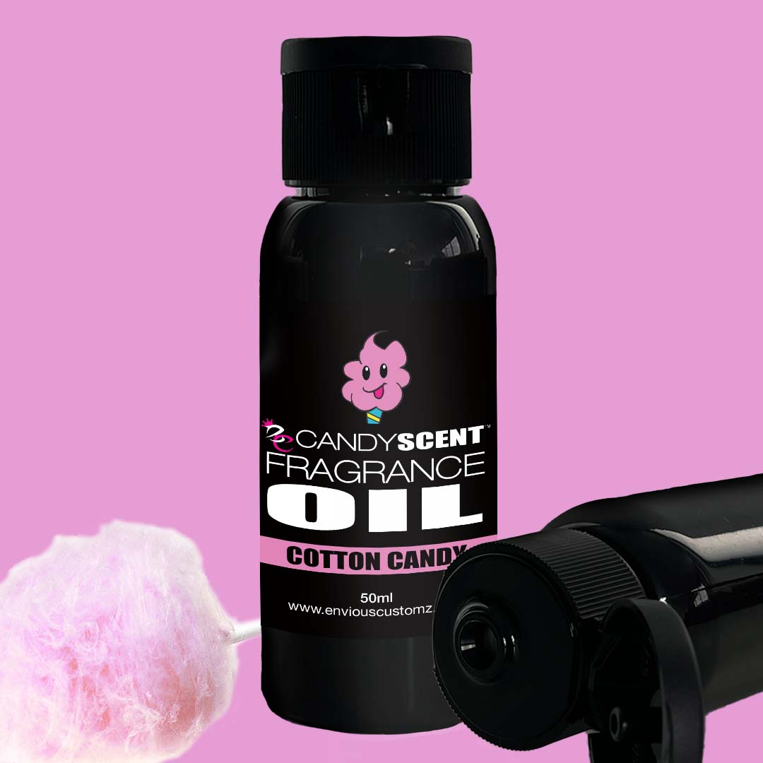 A black bottle labeled "CANDYSCENT™ Fragrance Oil" featuring a pink cartoon candy character stands out against a pink background. A smaller Fragrance Oil bottle lies horizontally nearby, with a piece of cotton candy placed to the left—ideal for your oil burner and home scent needs.