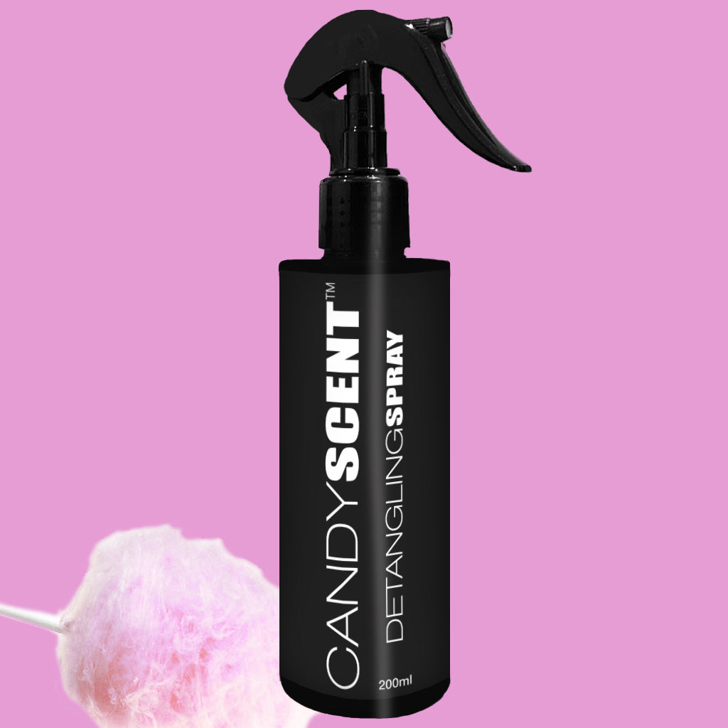 A black spray bottle labeled "CANDYSCENT™ Hair Detangling Spray" stands upright against a pink background. Enriched with jojoba oil, it promises smooth, tangle-free hair. In the lower left corner, there is a bundle of pink cotton candy on a stick.