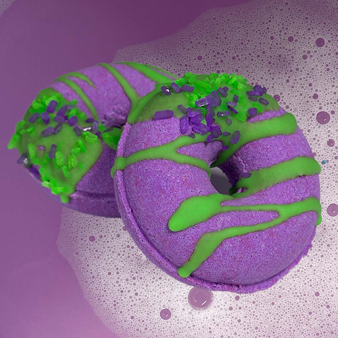 Two lively purple CANDYSCENT™ Bath Bombs, designed to look like donuts with green icing and sprinkles, float on a backdrop of bubbly water. The delightful scents combined with the playful colors create a visually captivating scene, all crafted from natural ingredients.