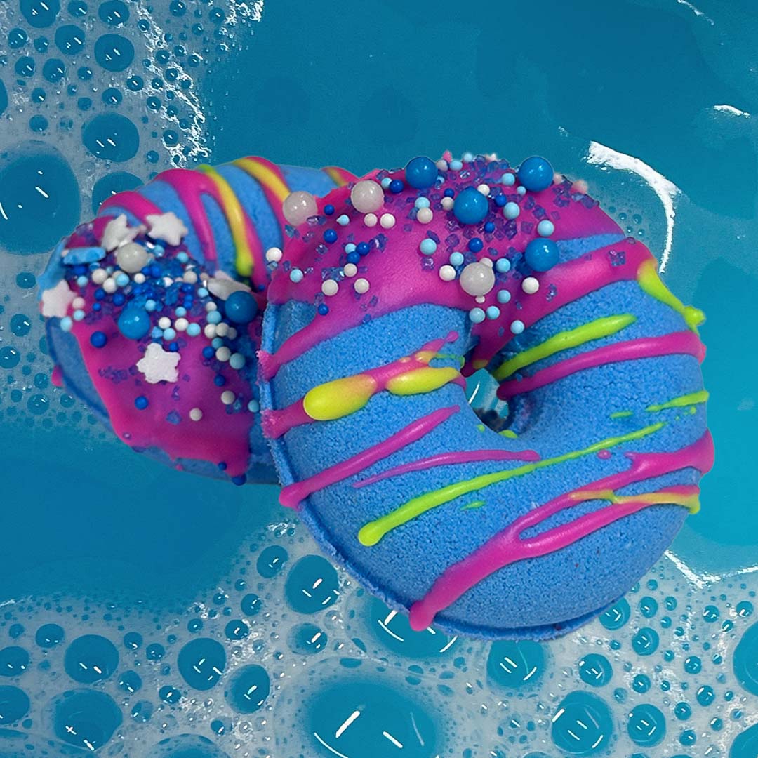 Two vibrant CANDYSCENT™ Bath Bombs float in the water. Predominantly blue with swirls of pink and purple, drizzled with neon green, they're decorated with colorful sprinkles and beads. Infused with sweet scents and natural ingredients, they create a whimsical and fun design perfect for any bath.