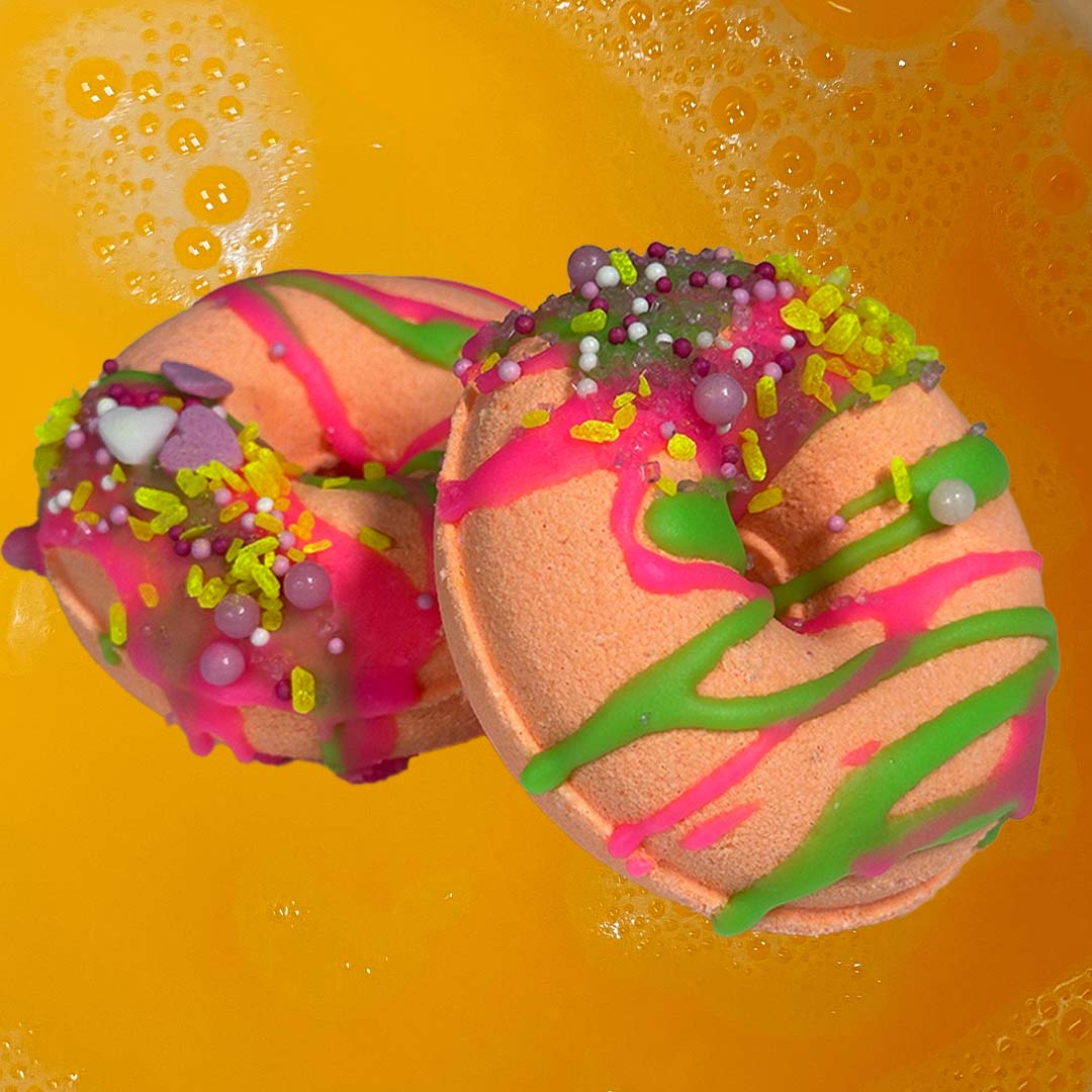 Two vibrant CANDYSCENT™ Bath Bombs, shaped like donuts, float in a bubbly orange liquid. Decorated with vivid pink, green, and purple drizzles and colorful sprinkles that resemble real frosting, these bath bombs not only look delightful but also release sweet scents from their natural ingredients.