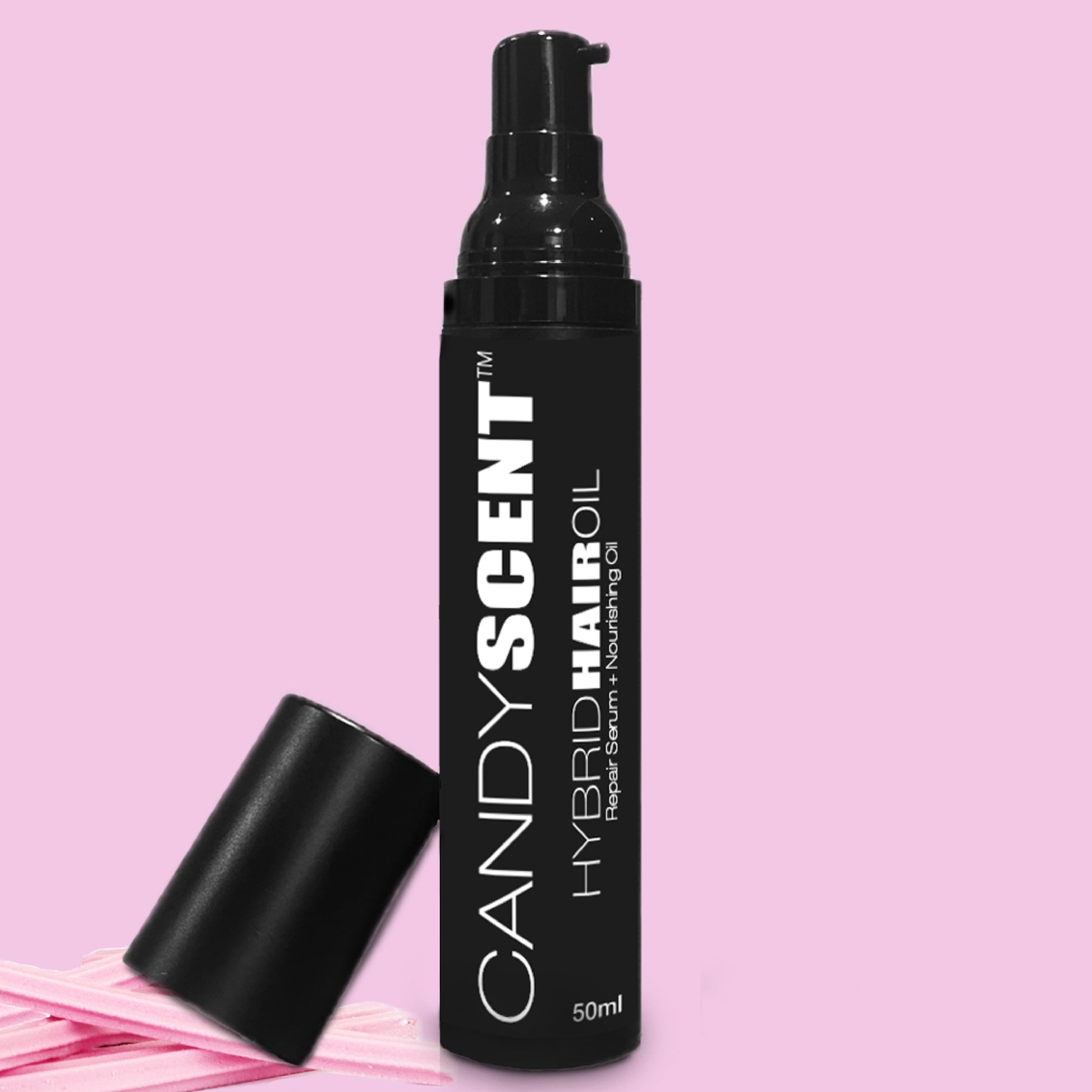 A black bottle of Hybrid Hair Oil by CANDYSCENT™ with a pump dispenser is positioned against a pink background. The bottle is labeled with the product name and details. The cap is removed and placed beside it, along with a couple of pink hair ties, showcasing the nourishing hair oil's allure.