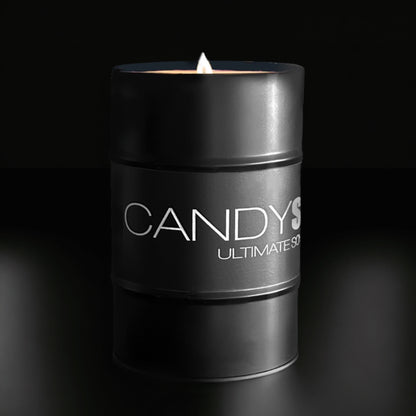 A black cylindrical candle from the CANDYSCENT™ brand, labeled "Soy Wax Candle Tin" and "Ultimate Soy," is lit and placed on a dark reflective surface. Made from soy wax, the flame emits a warm glow, creating a soft ambiance against the dark background, encapsulating the essence of CANDYSCENT™ candles.