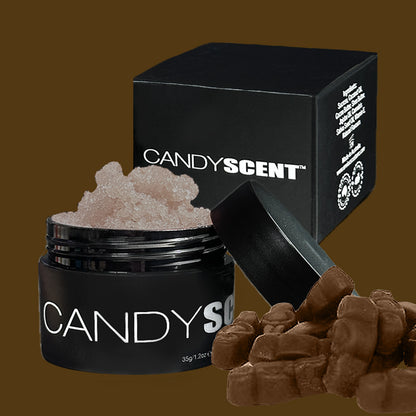 An open black container labeled "(DISCONTINUED CANDYSCENT™) Lip Sugar Scrubs" filled with a pinkish granulated substance promises a lip-smacking experience. The container's lid is slightly tilted to the side, next to a black box also displaying "CANDYSCENT™." In the foreground, several small chocolates add to the allure of this Lip Sugar Scrub for soft lips.