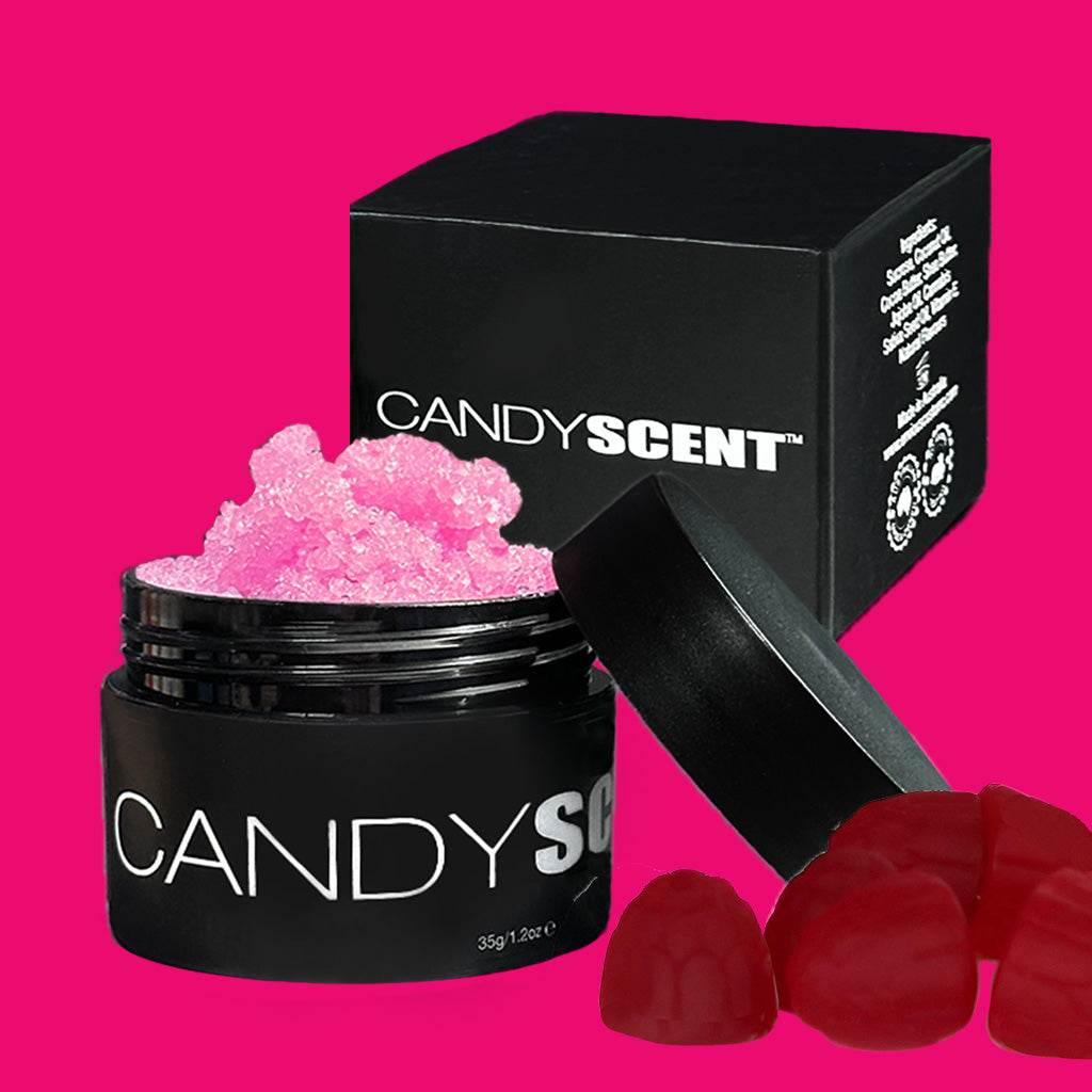 A black jar labeled "(DISCONTINUED CANDYSCENT™) Lip Sugar Scrubs," filled with pink granules, is open with its lid leaning against it. Behind the jar stands a matching black box. Red gummy candies are positioned in the lower right corner. The bright pink background enhances this tempting lip-smacking experience of our CANDYSCENT™ Lip Sugar Scrub for soft lips.